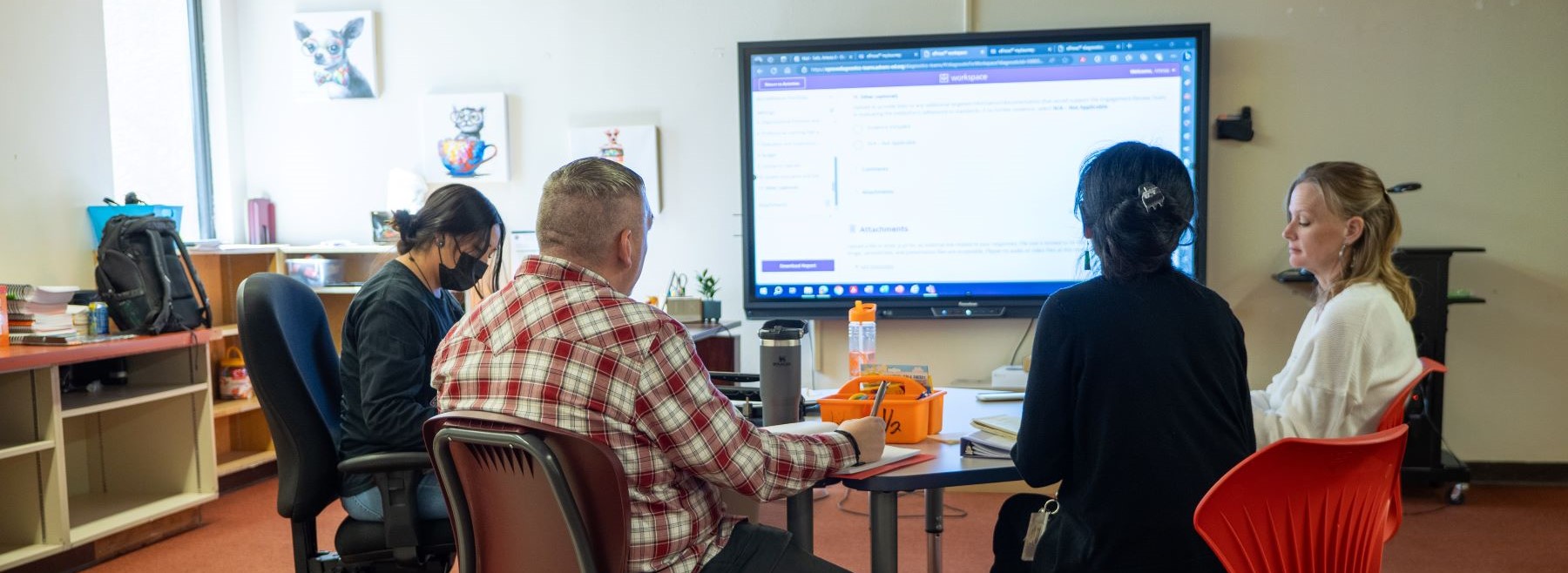 A group of school staff members seated around a table, engaged in discussion in front of a large presentation screen.